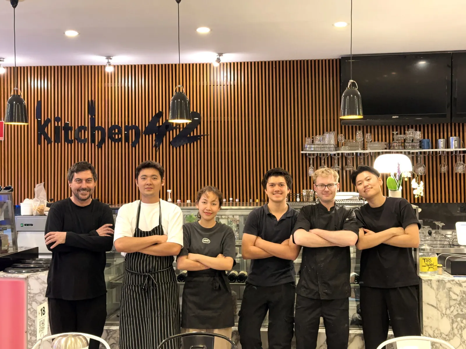 Six of the Kitchen 42 team standing in front of the timber wall and the Kitchen 42 sign